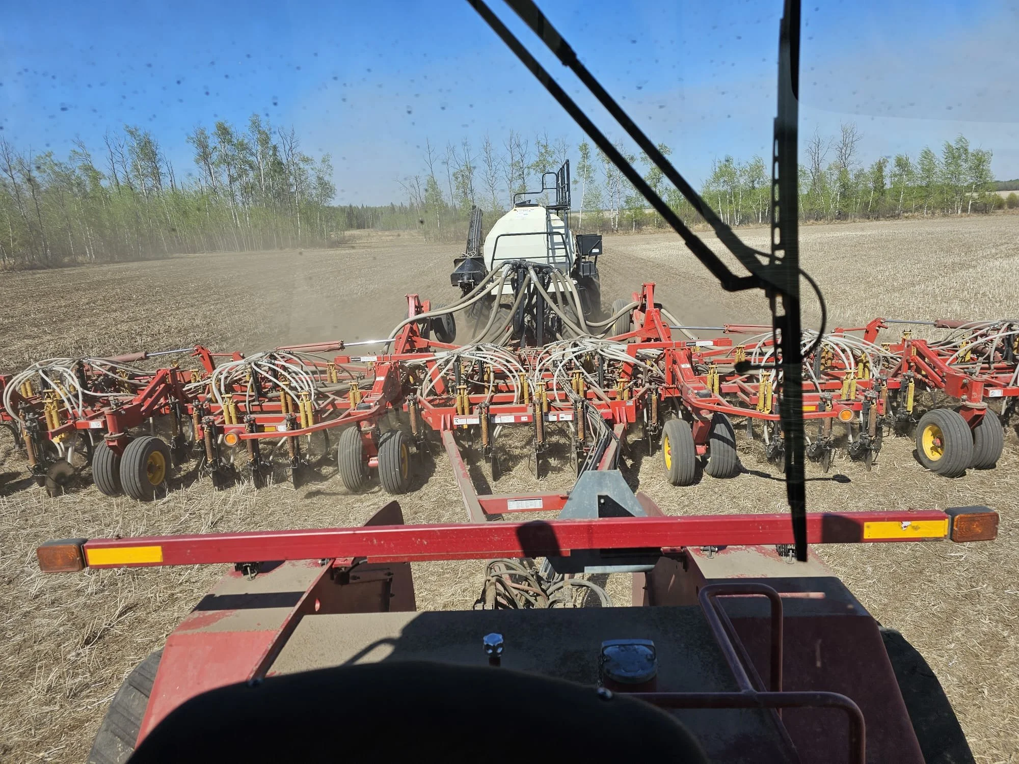 View from inside a tractor cab looking through the windshield at a large piece of red farm equipment tilling the soil in a field, with trees and blue sky in the background.