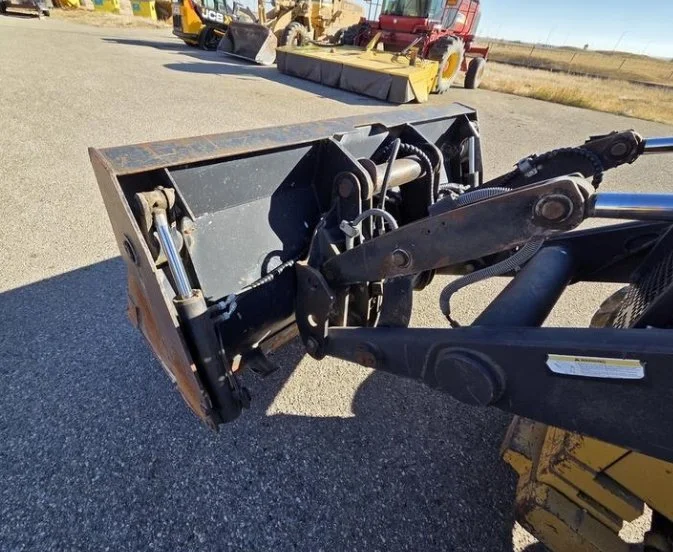Close-up of a front loader attachment on construction equipment, with a yellow bulldozer and other machinery in the background on a gravel surface.