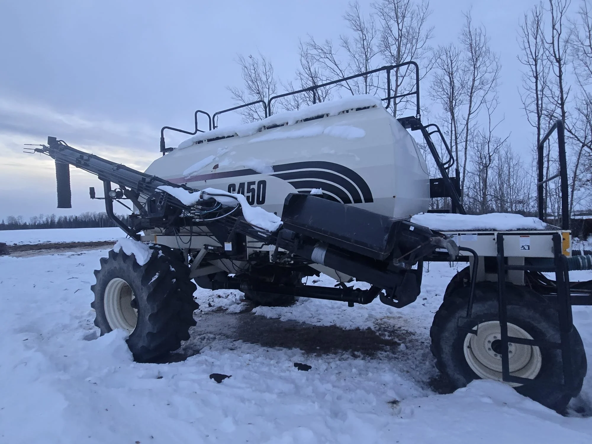 A large white agricultural or industrial sprayer vehicle with black and red stripes, situated in a snowy outdoor area with leafless trees in the background.