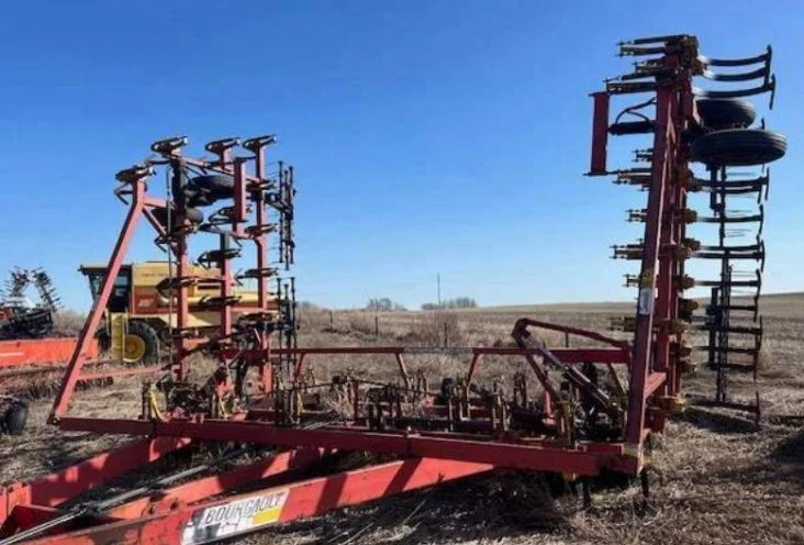 A large red agricultural seed planter with multiple rows of wheel and disc mechanisms, sitting in a field under a clear blue sky.
