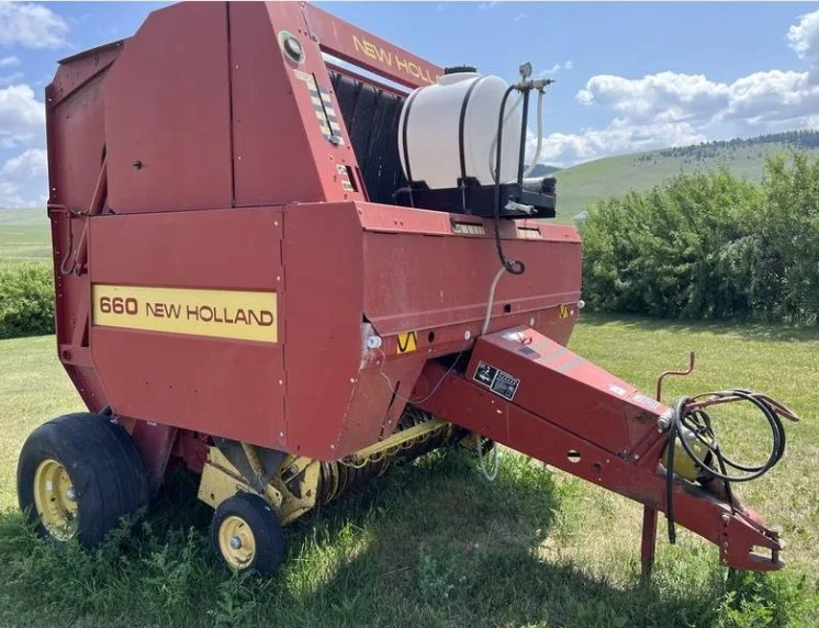 Red New Holland 660 baler in a grassy field with green trees and hills in the background under a partly cloudy sky.
