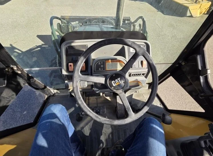 View from the driver's seat inside a yellow Caterpillar construction vehicle, showing the steering wheel, foot pedals, and control panel.