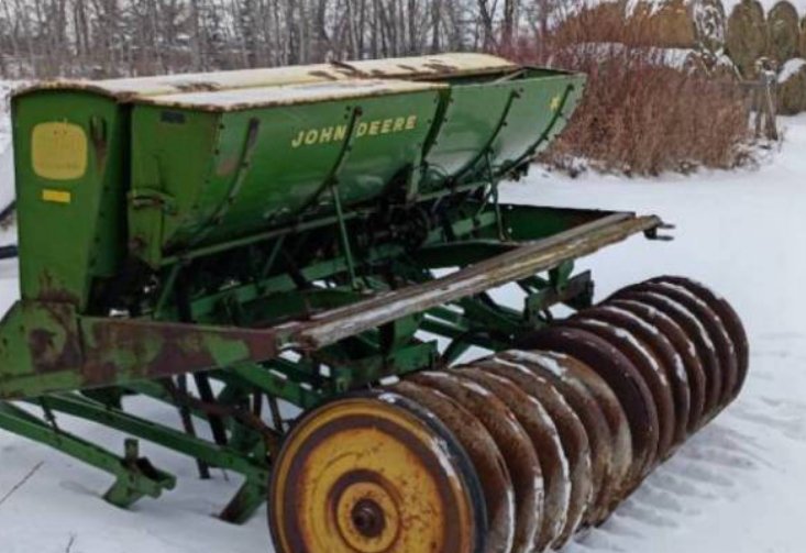 A John Deere farm seed or fertilizer planter machine on snow-covered ground with leafless trees in the background.