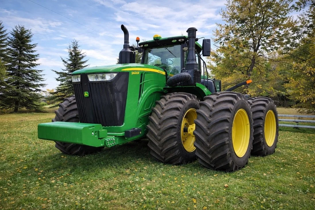 A large green John Deere tractor with six wheels on a grassy field with trees and a cloudy sky in the background.