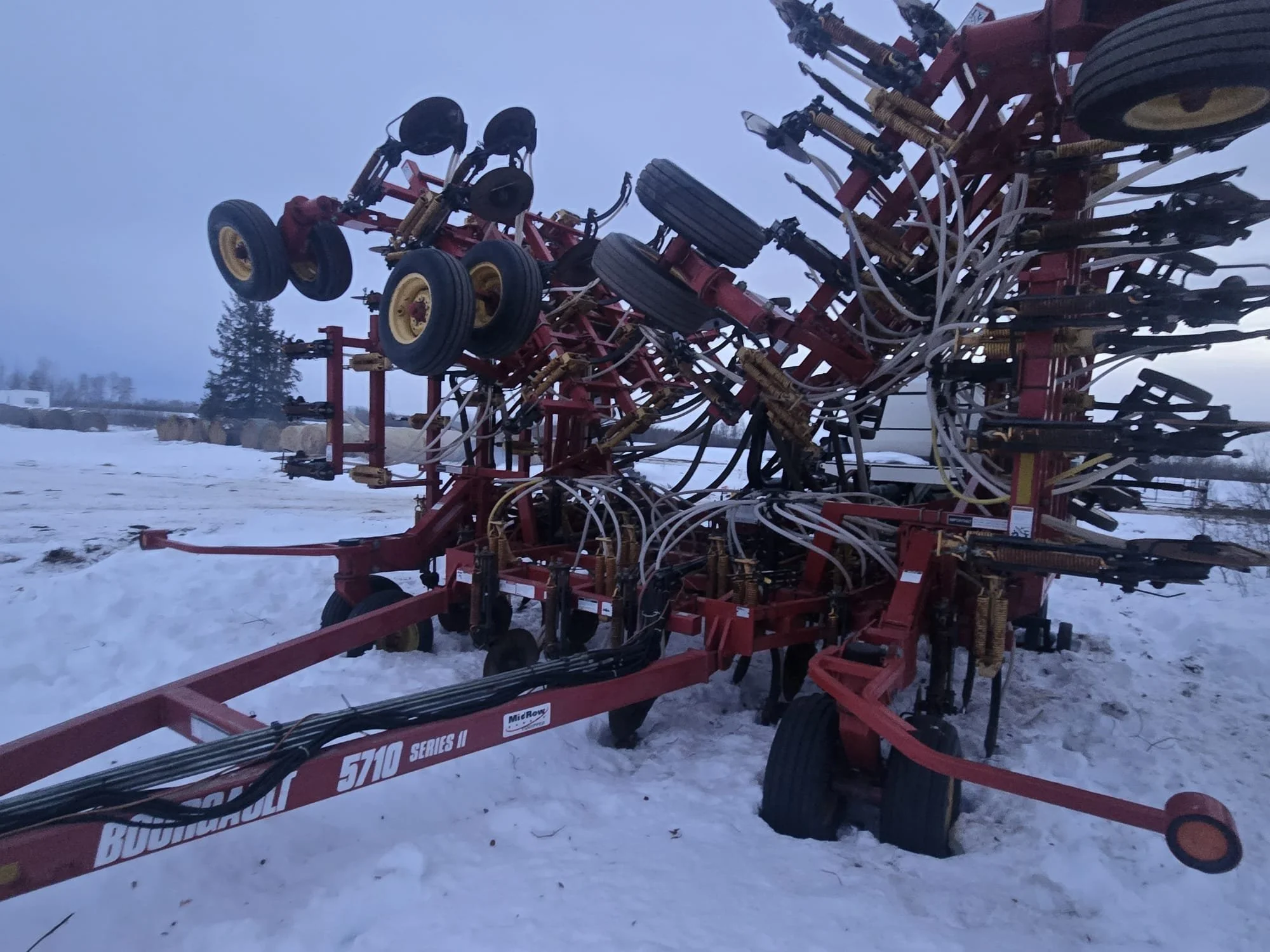 A large red farm machinery with multiple wheels, hydraulic hoses, and black discs, parked on snow-covered ground outdoors, with trees in the background.