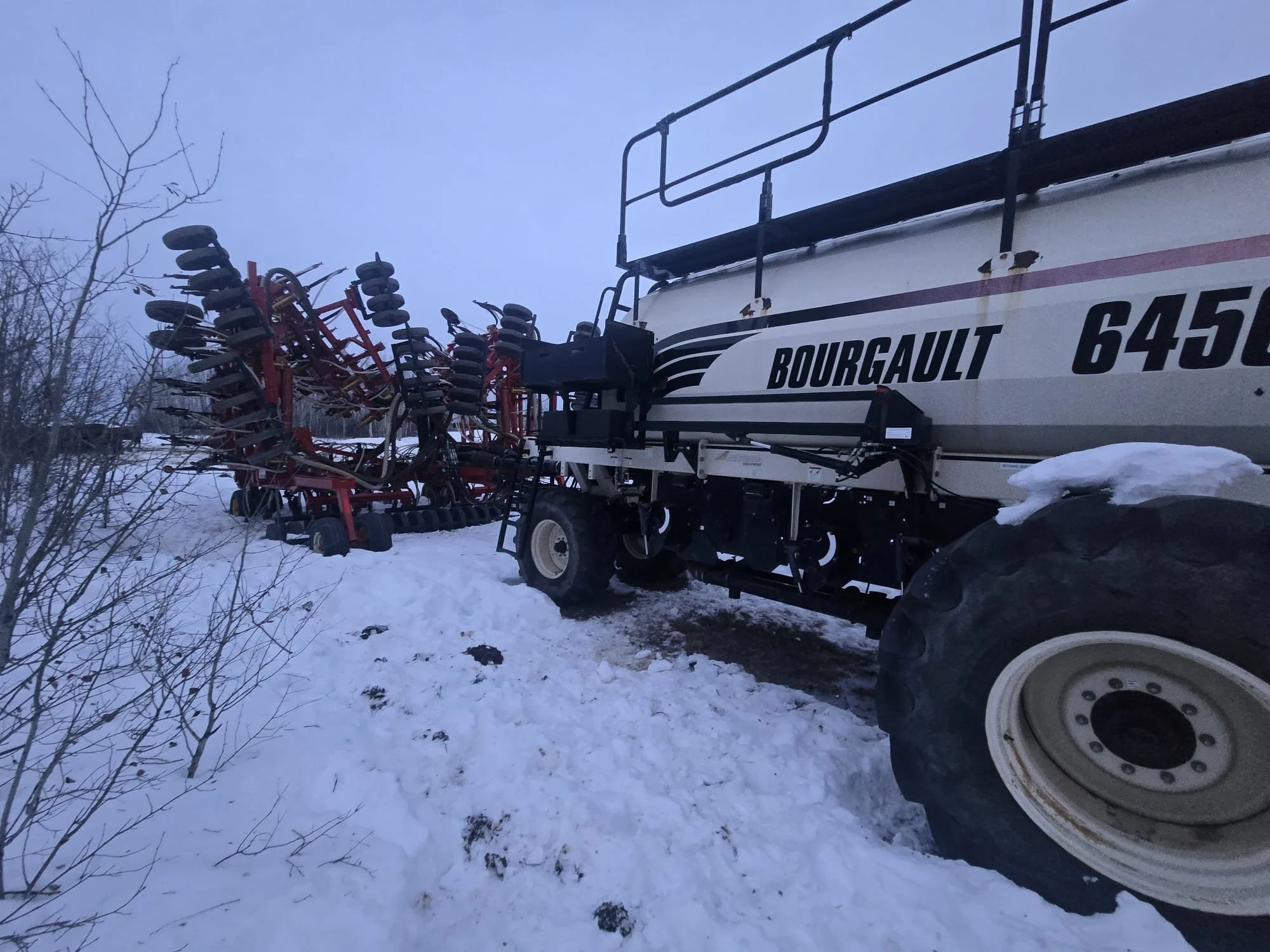 A large agricultural sprayer tractor, white with black lettering, is parked in snow with red equipment attached at the back.