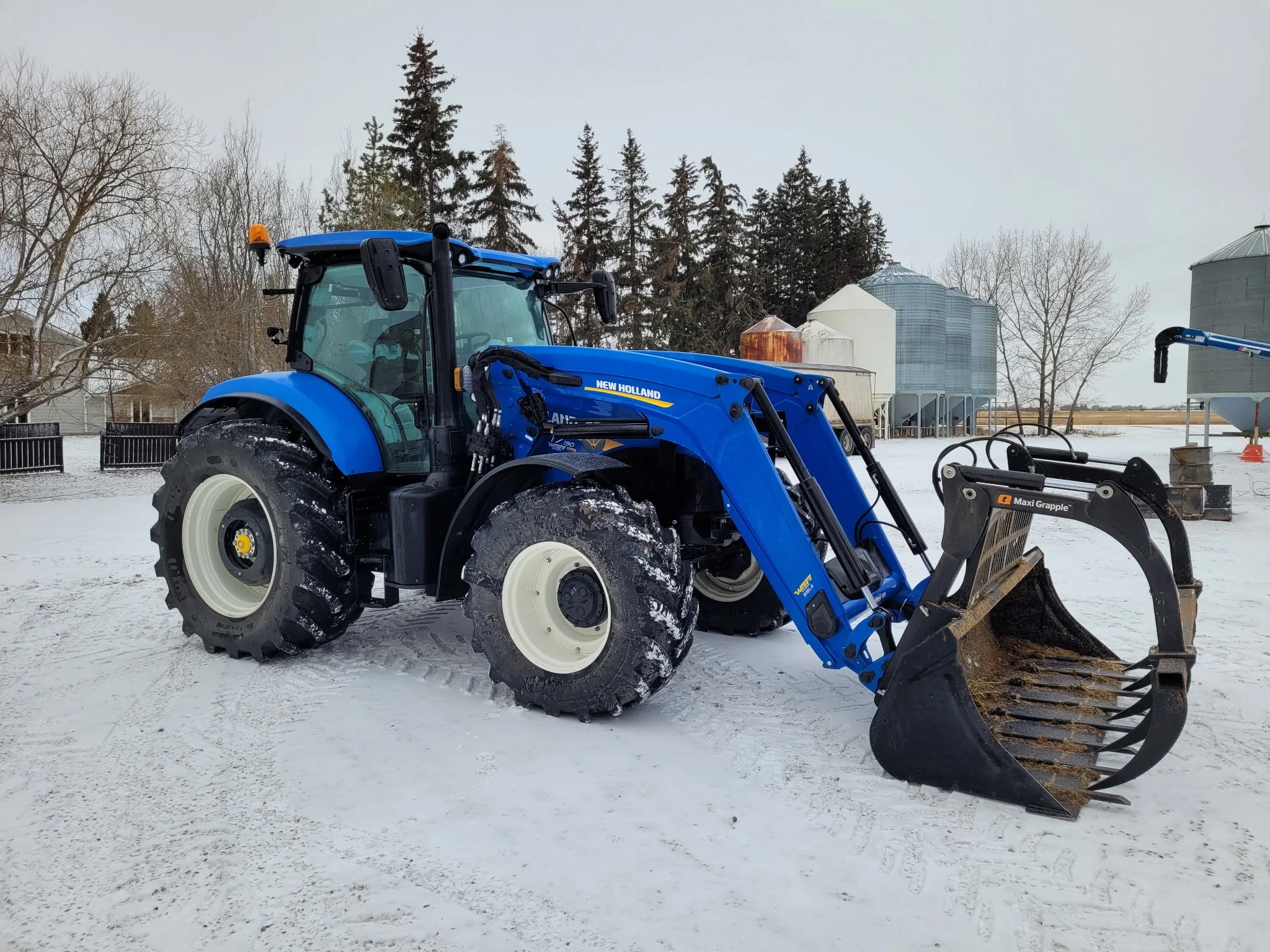 Blue New Holland compact tractor with front loader attachment on a snowy farm, with silos and trees in the background.