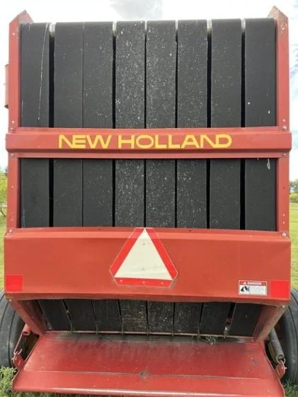 A red New Holland farm implement with black tires and vertical black weight blocks, featuring a reflective triangle safety sign on the back.