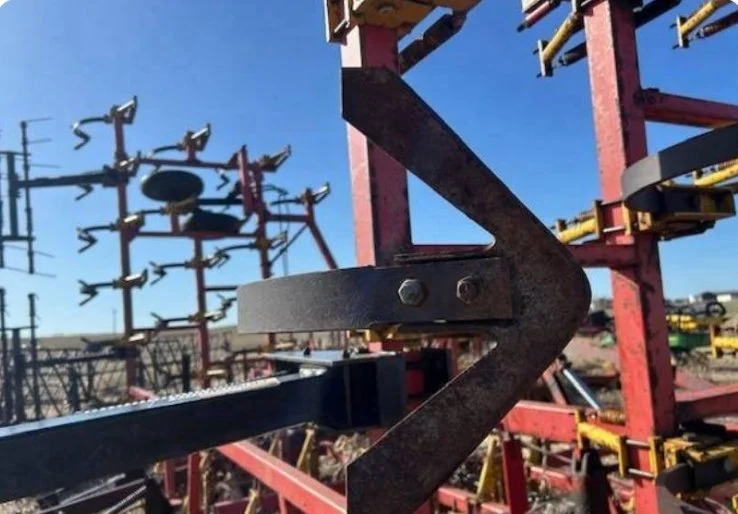 Close-up of a rusty metal tractor part with a zigzag shape, part of red farm equipment in the background under a clear blue sky.