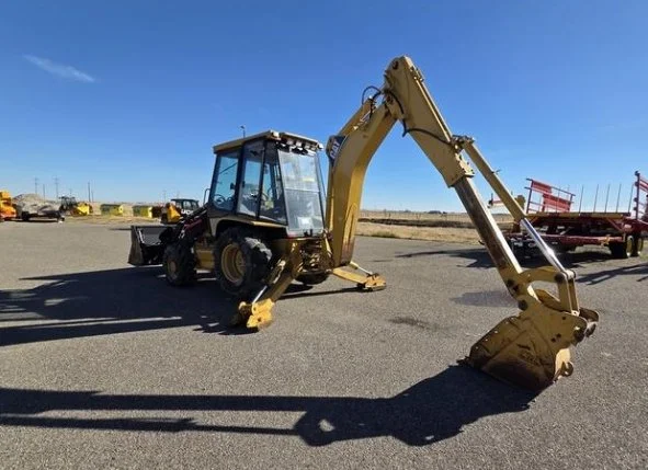 Yellow backhoe with a bucket attachment parked on asphalt in an open lot with other construction vehicles in the background.
