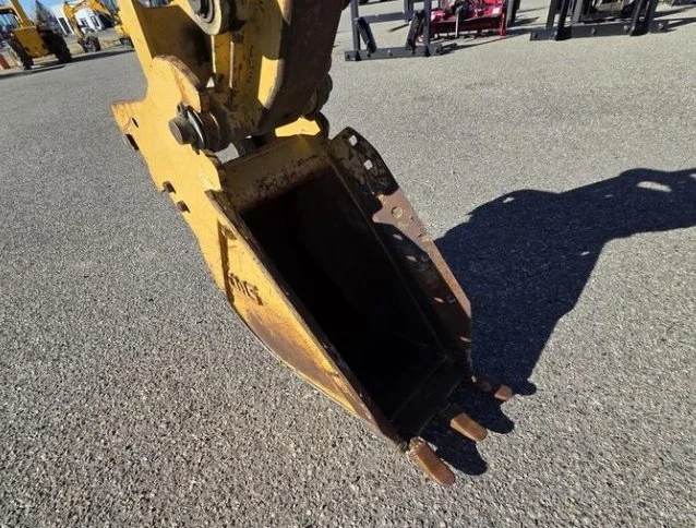 Close-up of a yellow excavator bucket on a gravel surface, with shadows casting on the ground.