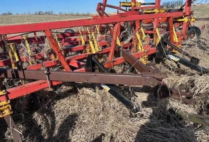 A red farm implement with yellow and black mechanical parts, fitted with multiple wheels, lying on dry grass and soil in an open field.