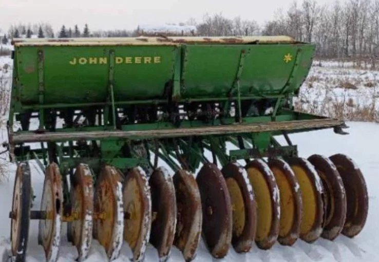 Green John Deere seed planter on snow-covered ground with trees in the background.