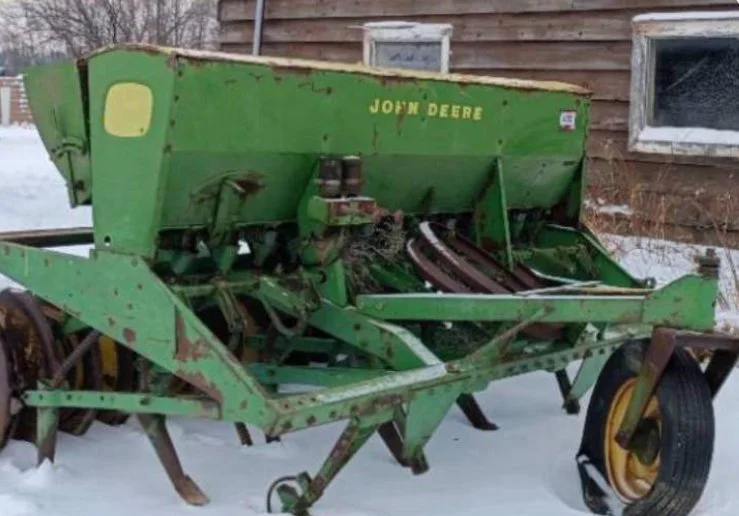 Old green John Deere farm equipment on snow, with a wooden building and windows in background.