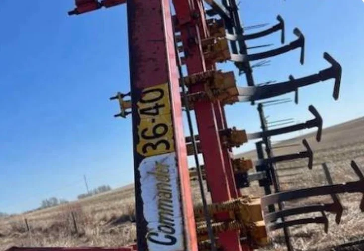 A red farm machinery implement with curved tines and yellow warning decals, standing outdoors in a field under a clear blue sky.