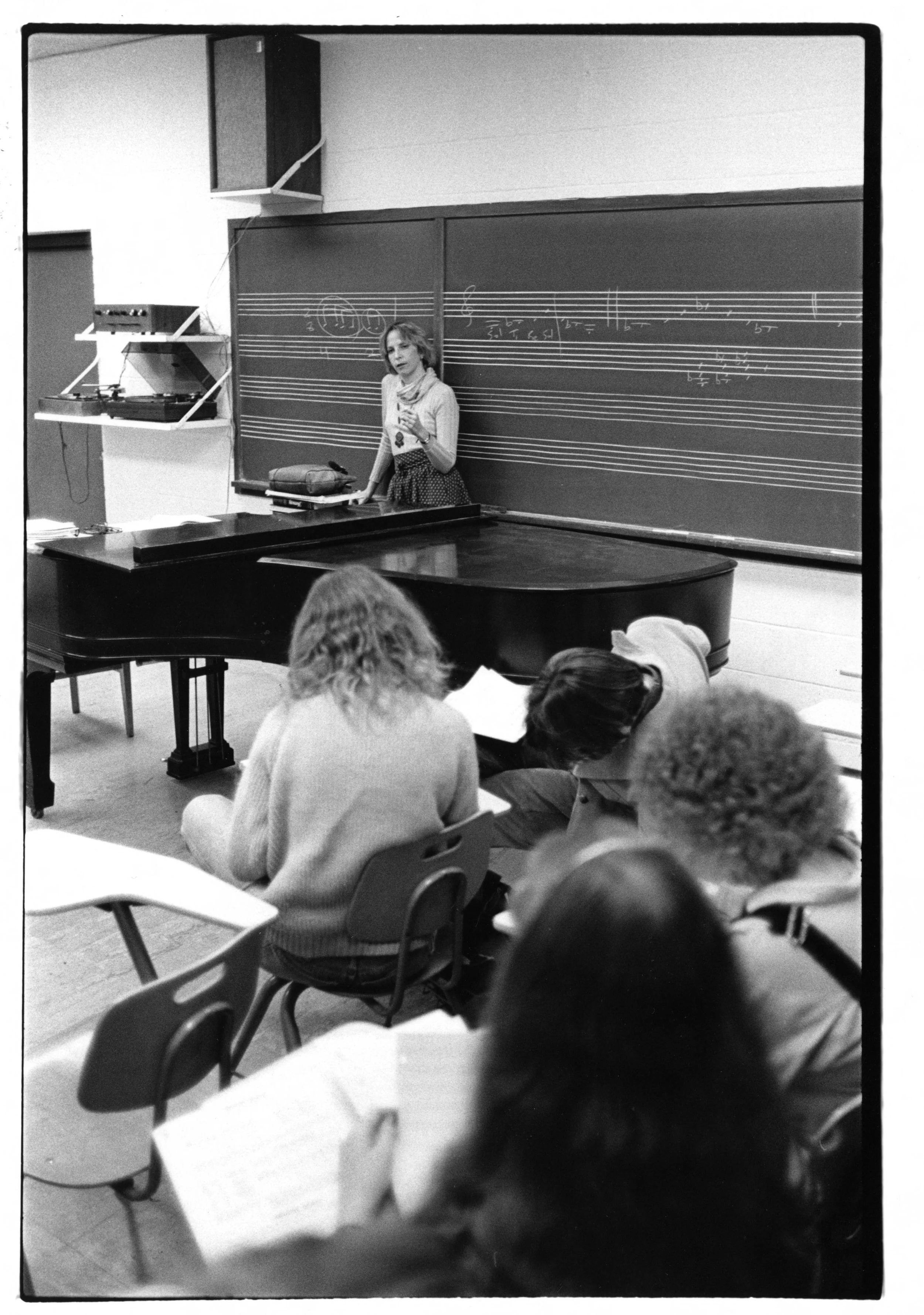 A classroom scene with a woman, Faye-Ellen Silverman, standing at a blackboard, which has musical notation and notes written on it. Students are seated at desks, taking notes and listening to her.