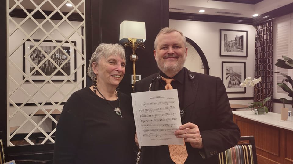 A smiling Faye-Ellen Silverman and smiling man holding sheet music in a warmly decorated room with framed pictures, a lamp, potted plants, and striped chairs.