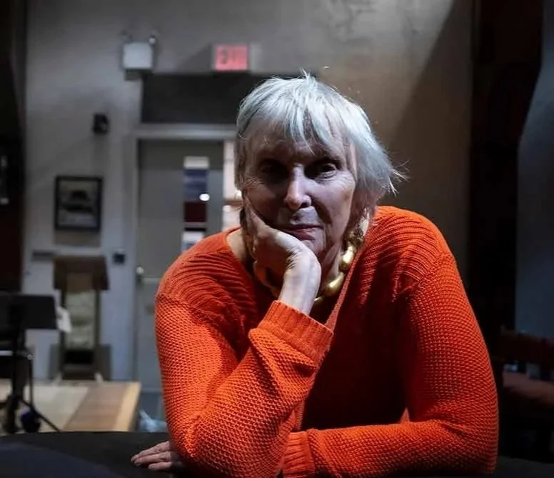 Portrait of Faye-Ellen Silverman with short gray hair, wearing an orange sweater and a gold necklace, resting her chin on her hand while sitting at a table in a dimly lit indoor space.