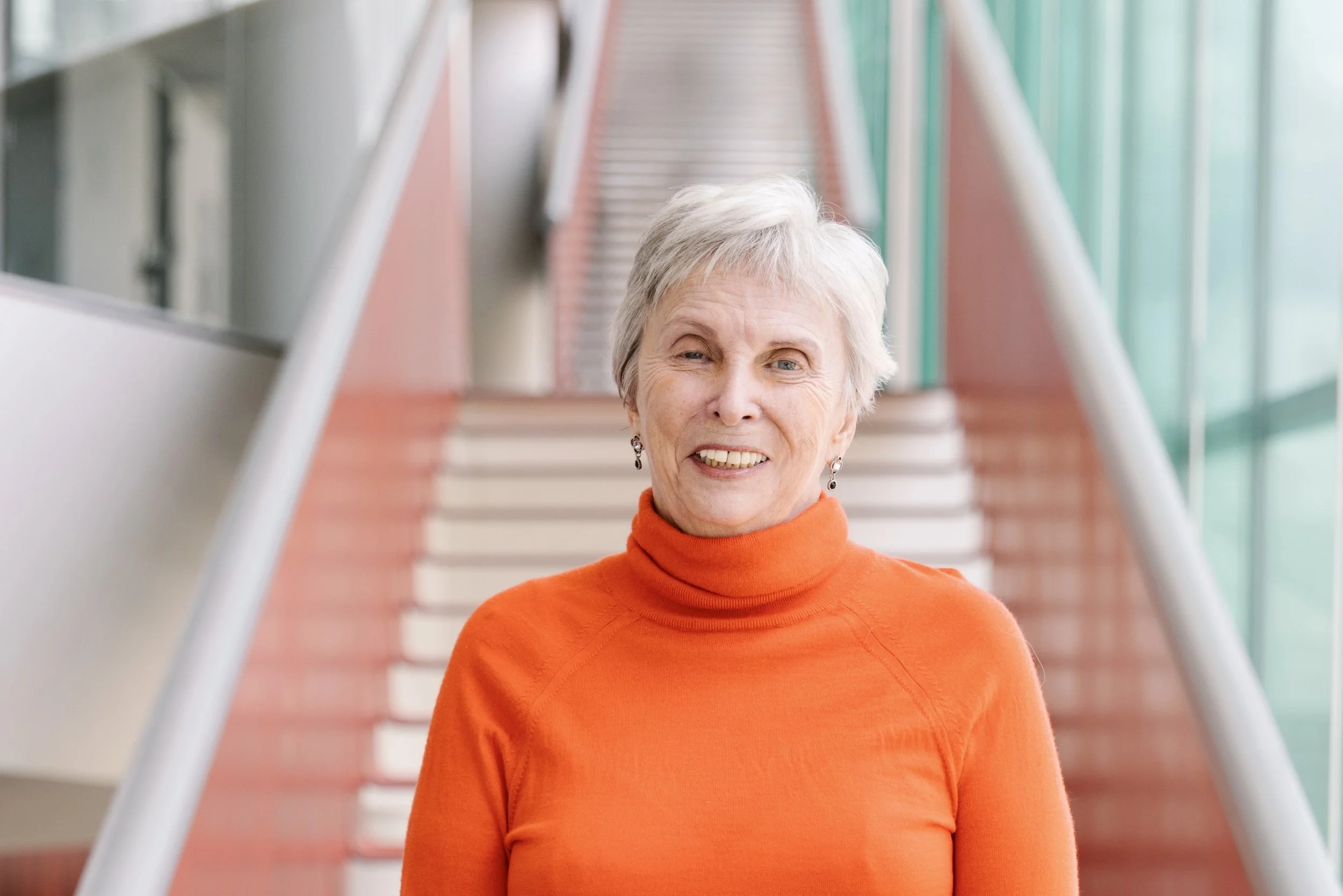 Headshot of Faye-Ellen Silverman with short gray hair wearing an orange turtleneck sweater standing on a staircase inside a modern building with glass walls.