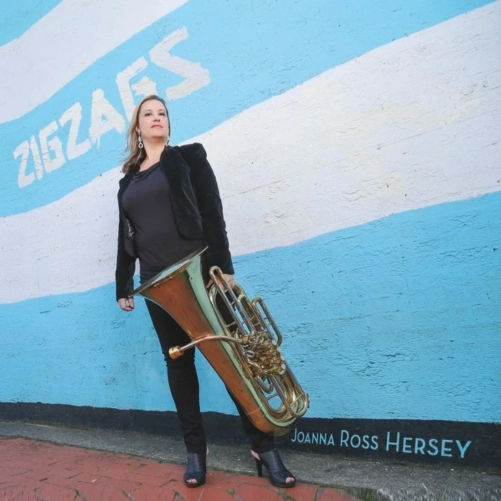 Cover album featuring a woman standing outdoors next to a wall with the word 'ZIGZAGS' painted on it. She is holding a large tuba and wearing a black jacket, dark pants, and high-heeled shoes.