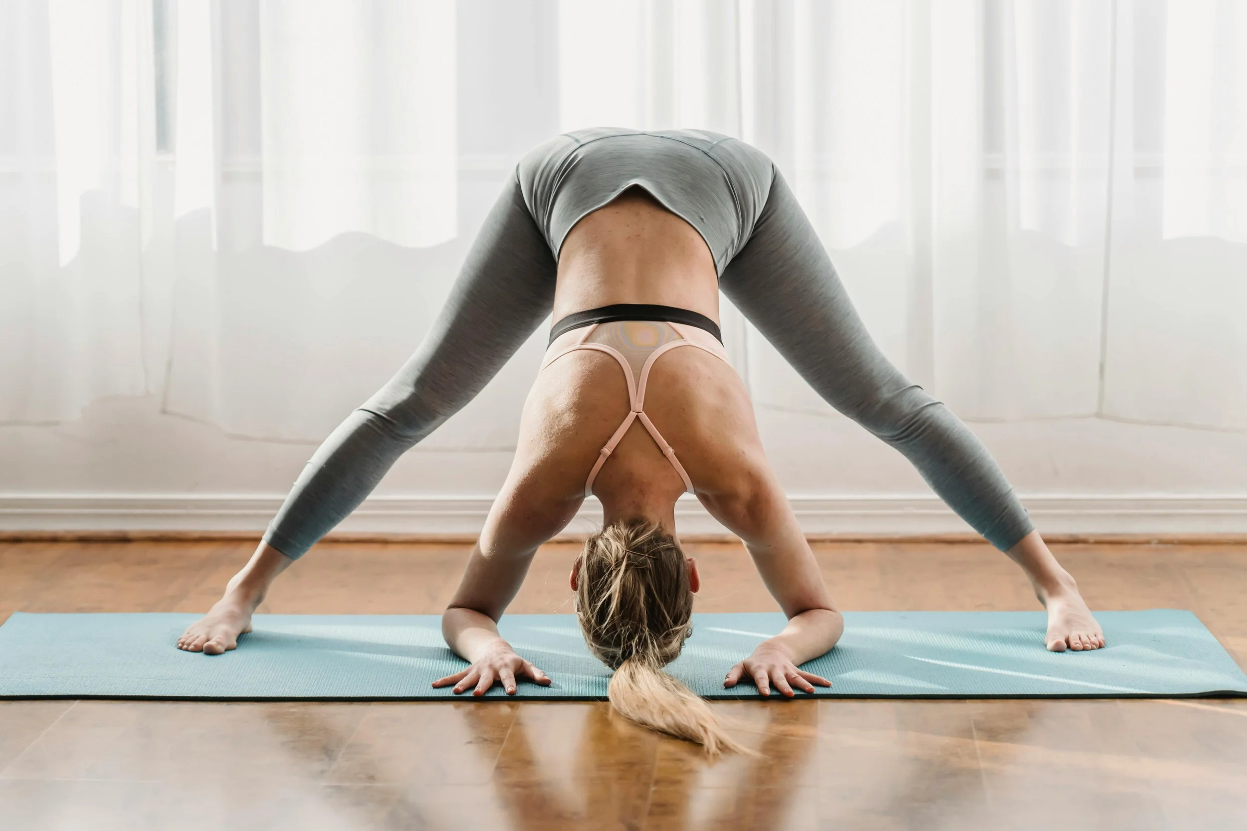 A woman doing a downward dog yoga pose on a light blue mat in a bright room with white curtains.