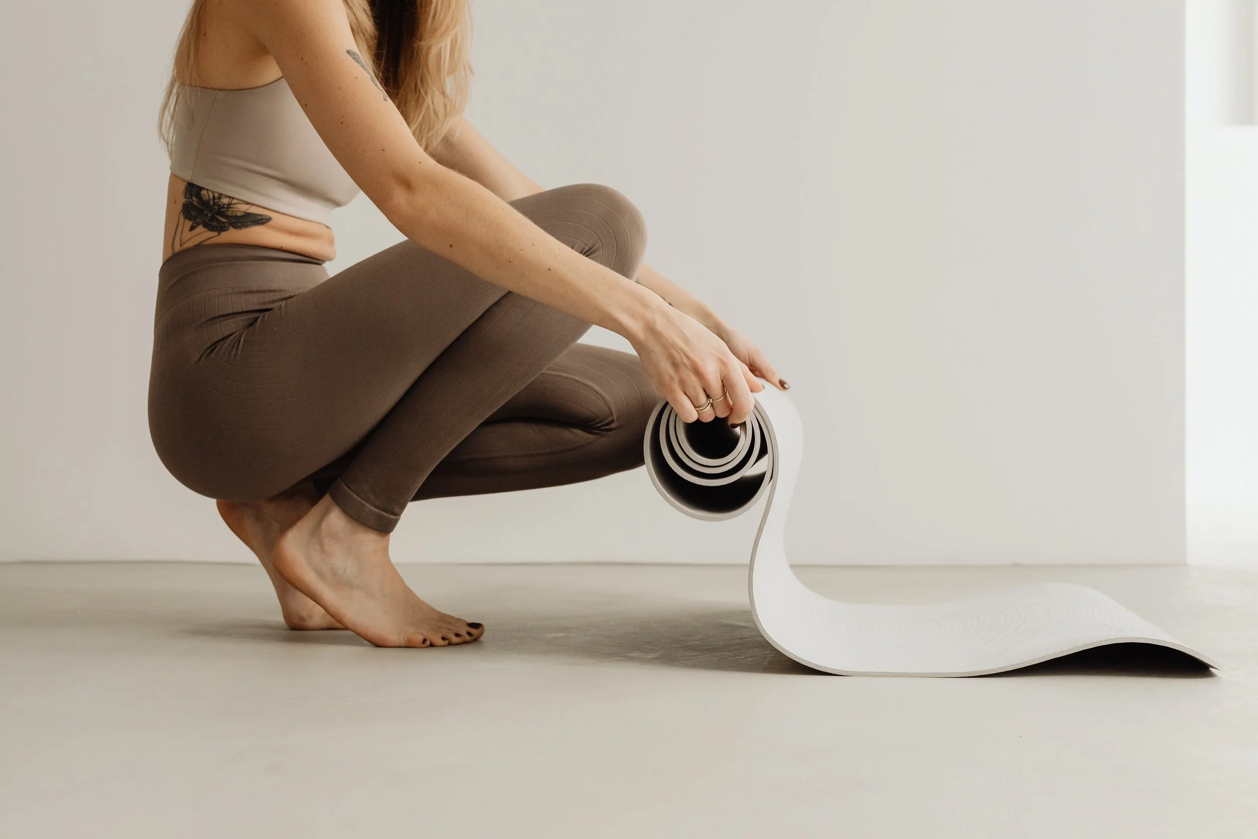 A woman with red hair and tattoos is crouching on the floor, holding a rolled-up yoga mat, with her feet bare on a plain, light-colored surface near a white wall.