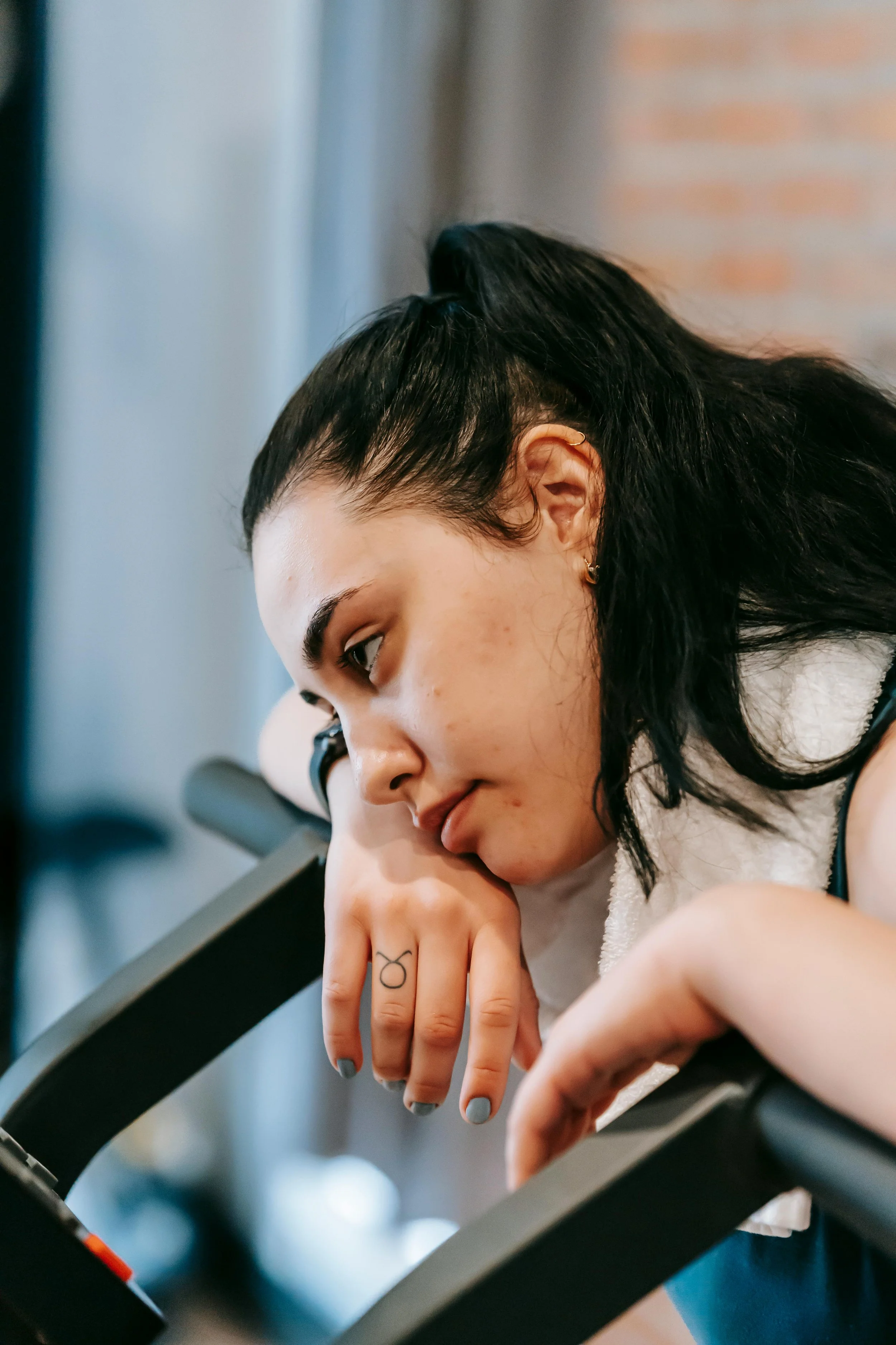 A young woman with dark hair tied back, wearing a small earring, is resting her chin on her hand while looking downward. She has a tattoo on her finger and appears to be sitting by a window with natural light.