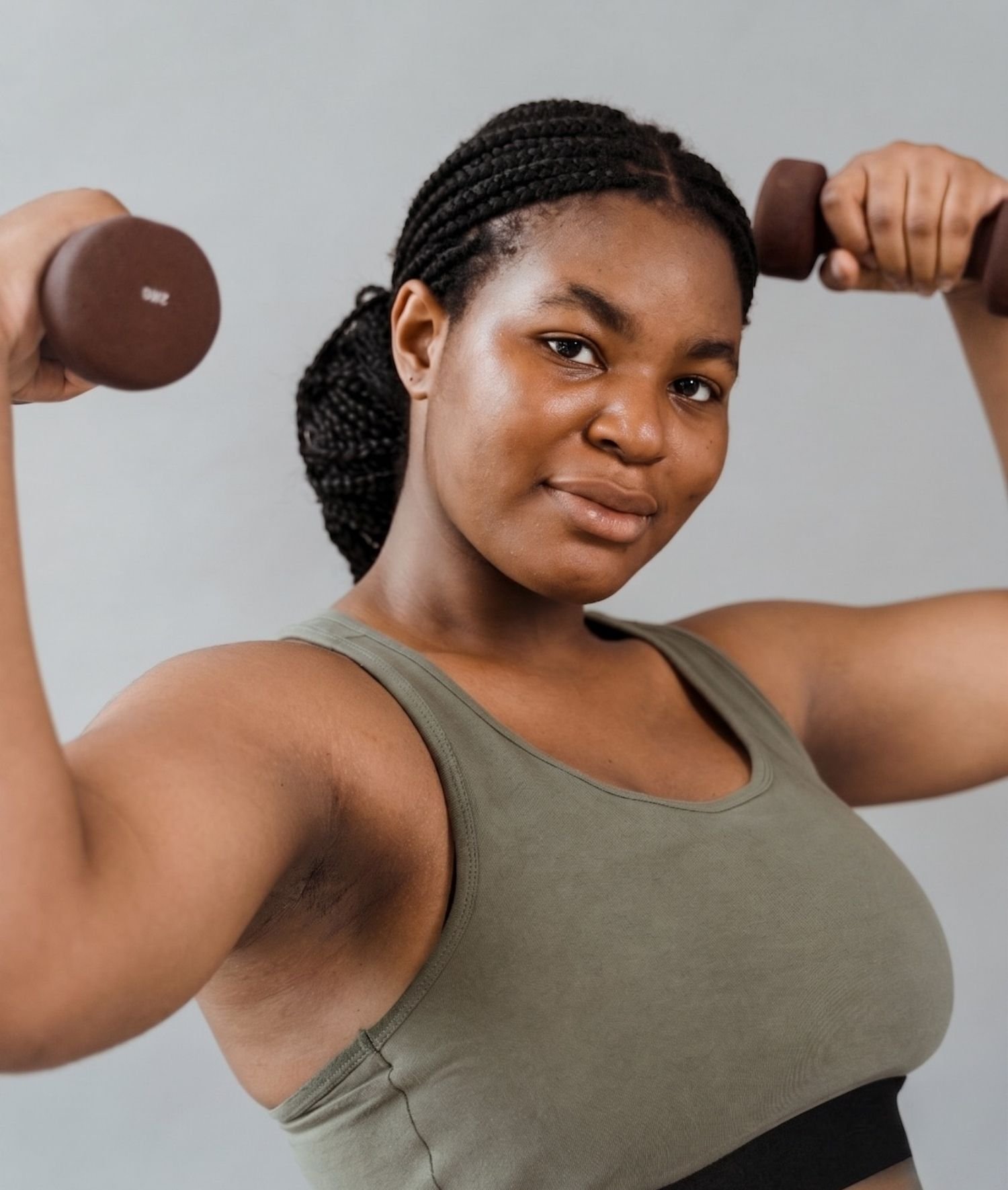 A woman with dark braided hair lifting brown dumbbells in a workout. She is wearing a sleeveless olive green top and has a neutral expression. The background is plain and light-colored.