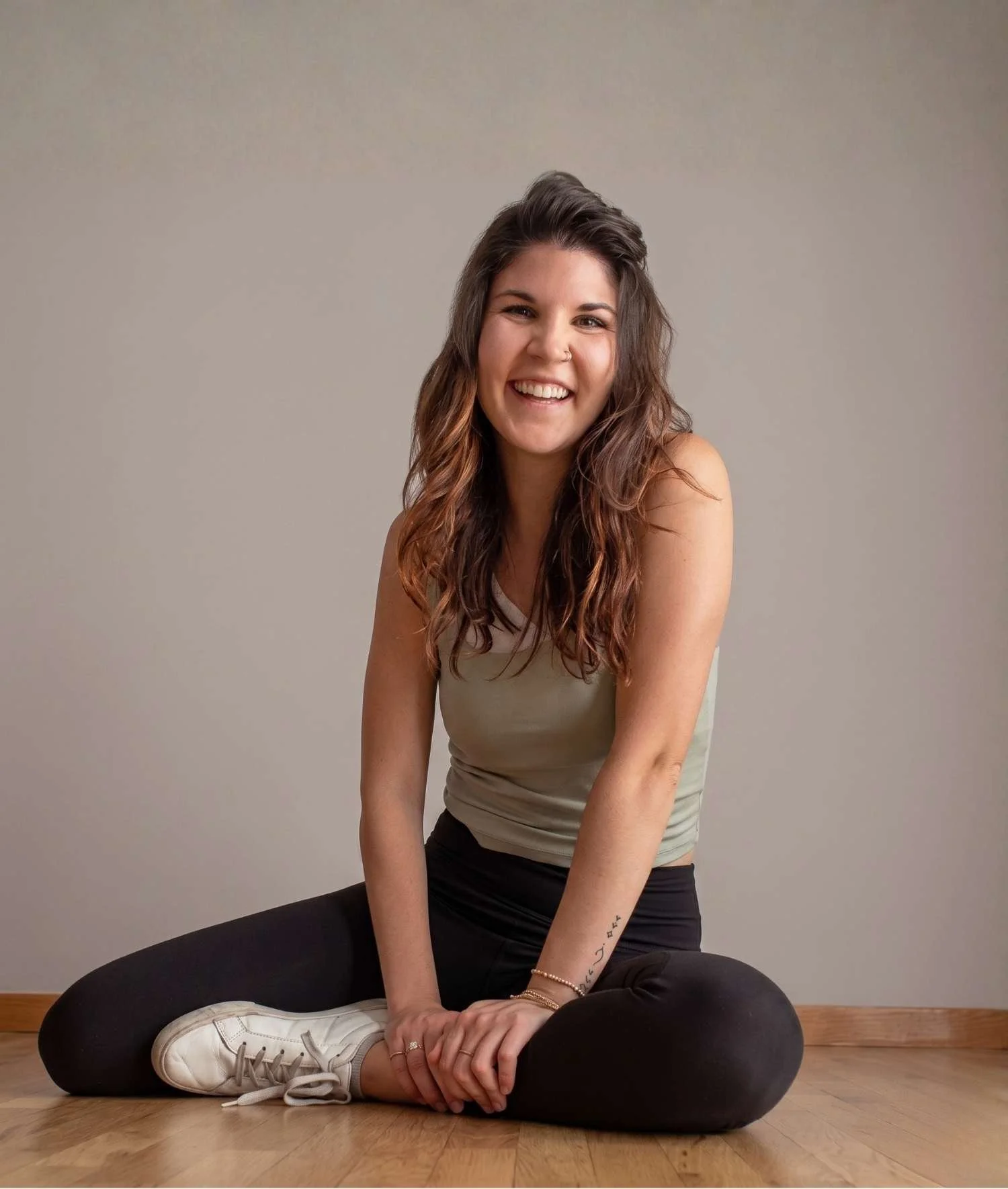 Woman with long brown wavy hair, wearing a light-colored tank top, black pants, and white sneakers, sitting cross-legged on a wooden floor, smiling and looking at the camera, with a neutral background.