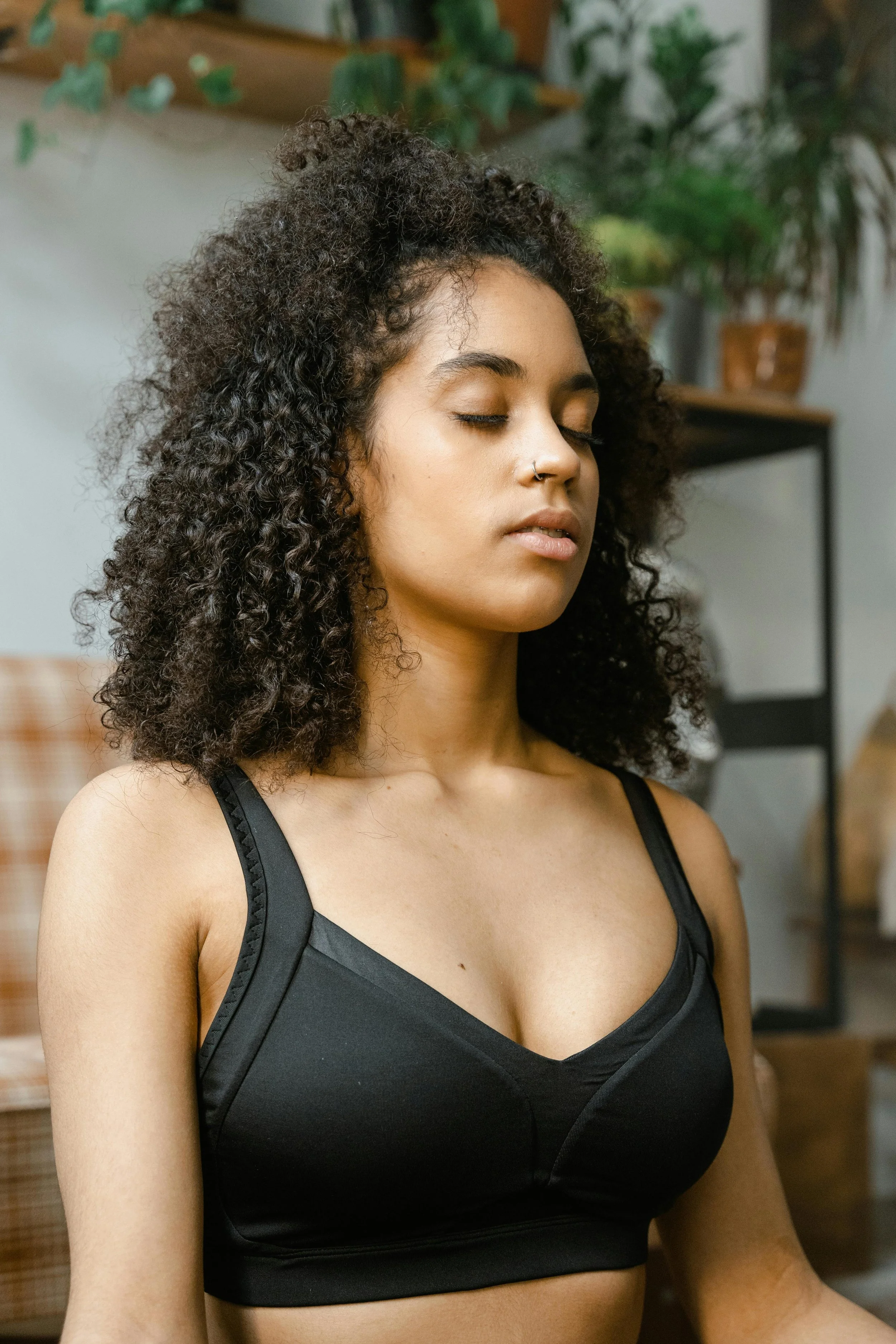 A woman with curly dark hair, closed eyes, and a nose ring, wearing a black top with lace straps, sitting in a room with plants in the background.