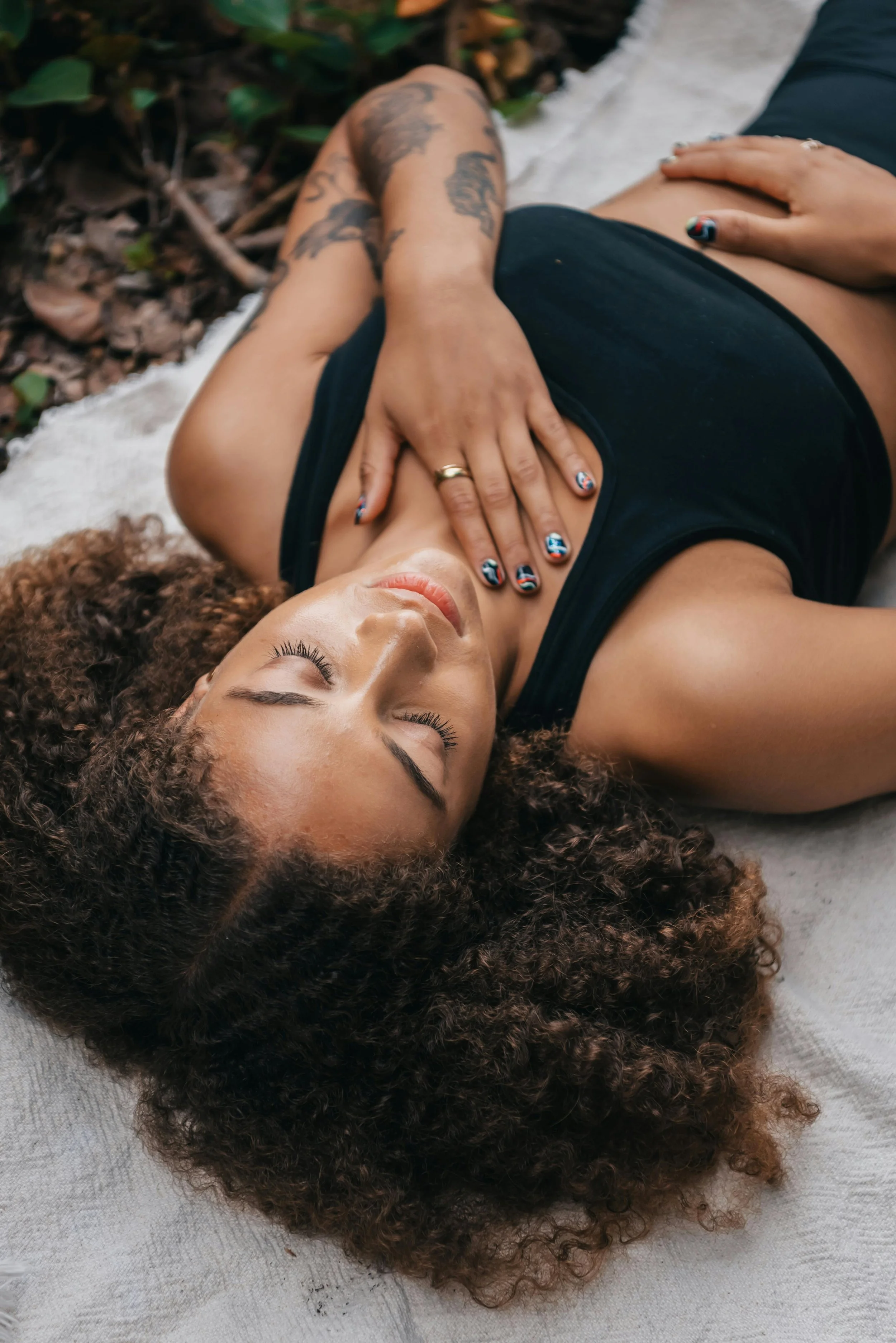 A woman with curly hair lying down on a gray fabric surface, eyes closed, wearing a black sleeveless top. One hand is resting on her chest, and the other is visible on her stomach.