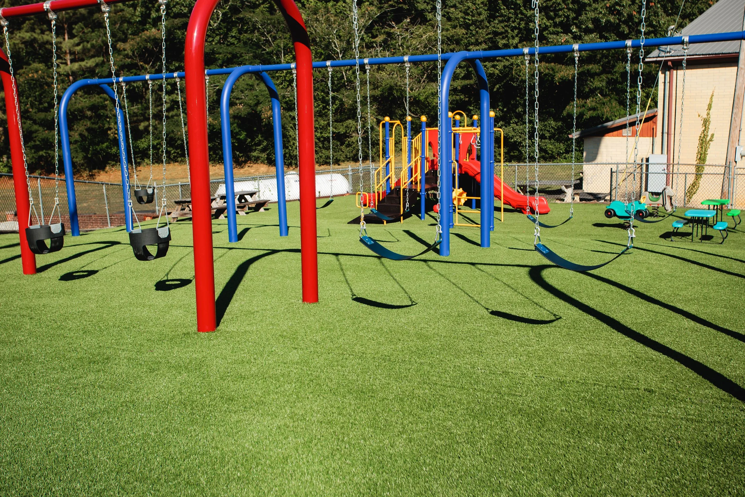 Colorful playground with red and blue swings, a slide, and small ride-on cars on green turf, surrounded by a fence and trees in the background.