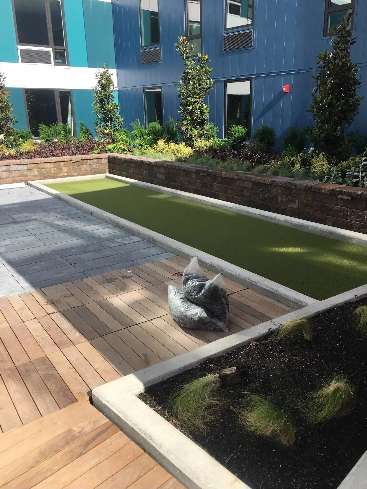 Urban outdoor courtyard with a small rectangular grassy area, wooden and stone paving, a brick wall, potted plants, and a modern blue building in the background.