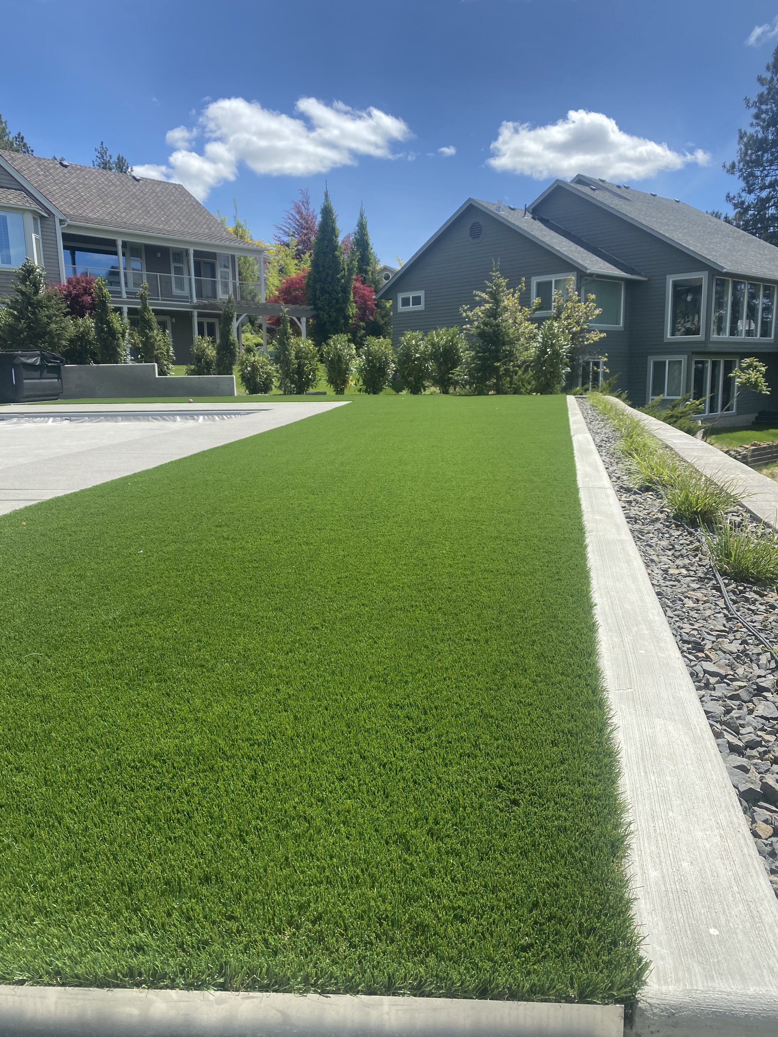 Manicured green lawn with concrete border, side of house with townhouses and trees under a blue sky with clouds
