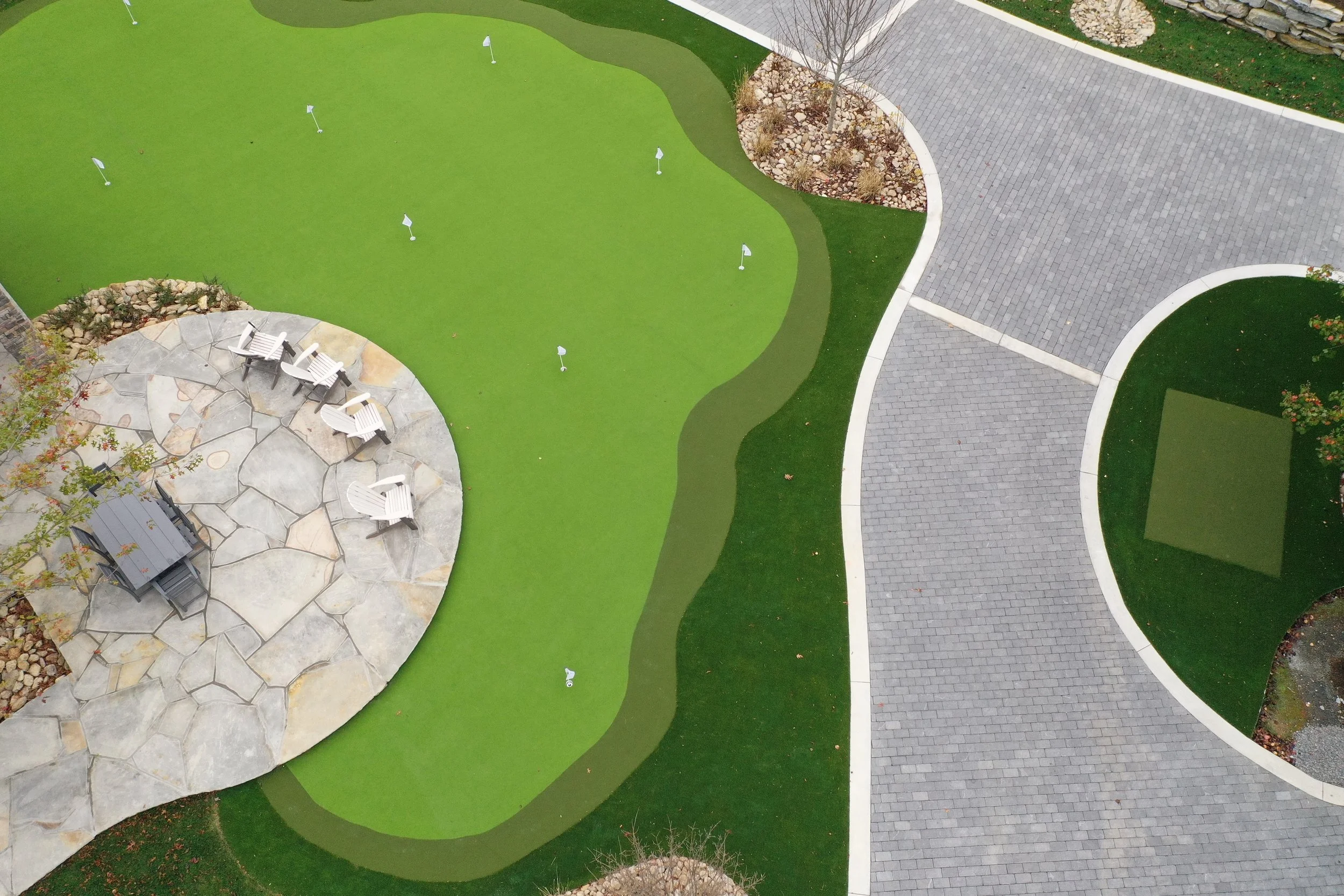 A landscaped backyard with a circular stone patio area with chairs and tables, a putting green, and a paved walkway with curved edges.