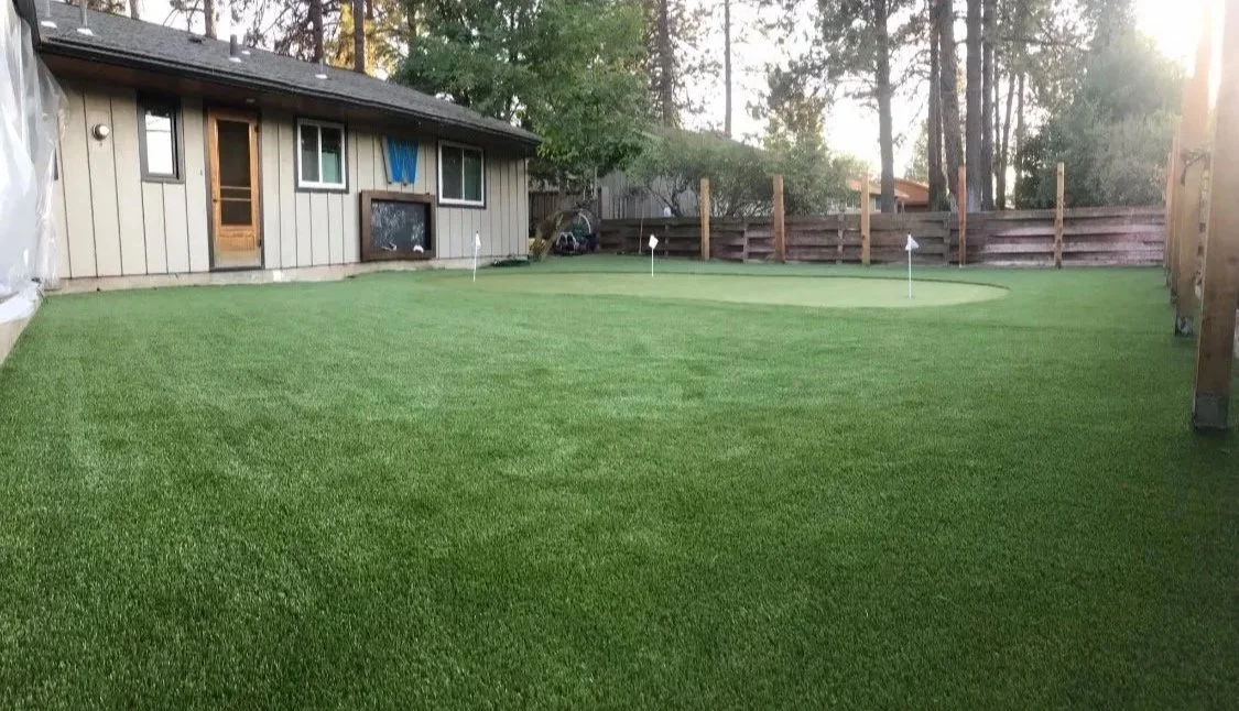 Backyard with artificial grass, small golf putting green, and flags, fenced with wooden privacy fence, surrounded by trees.