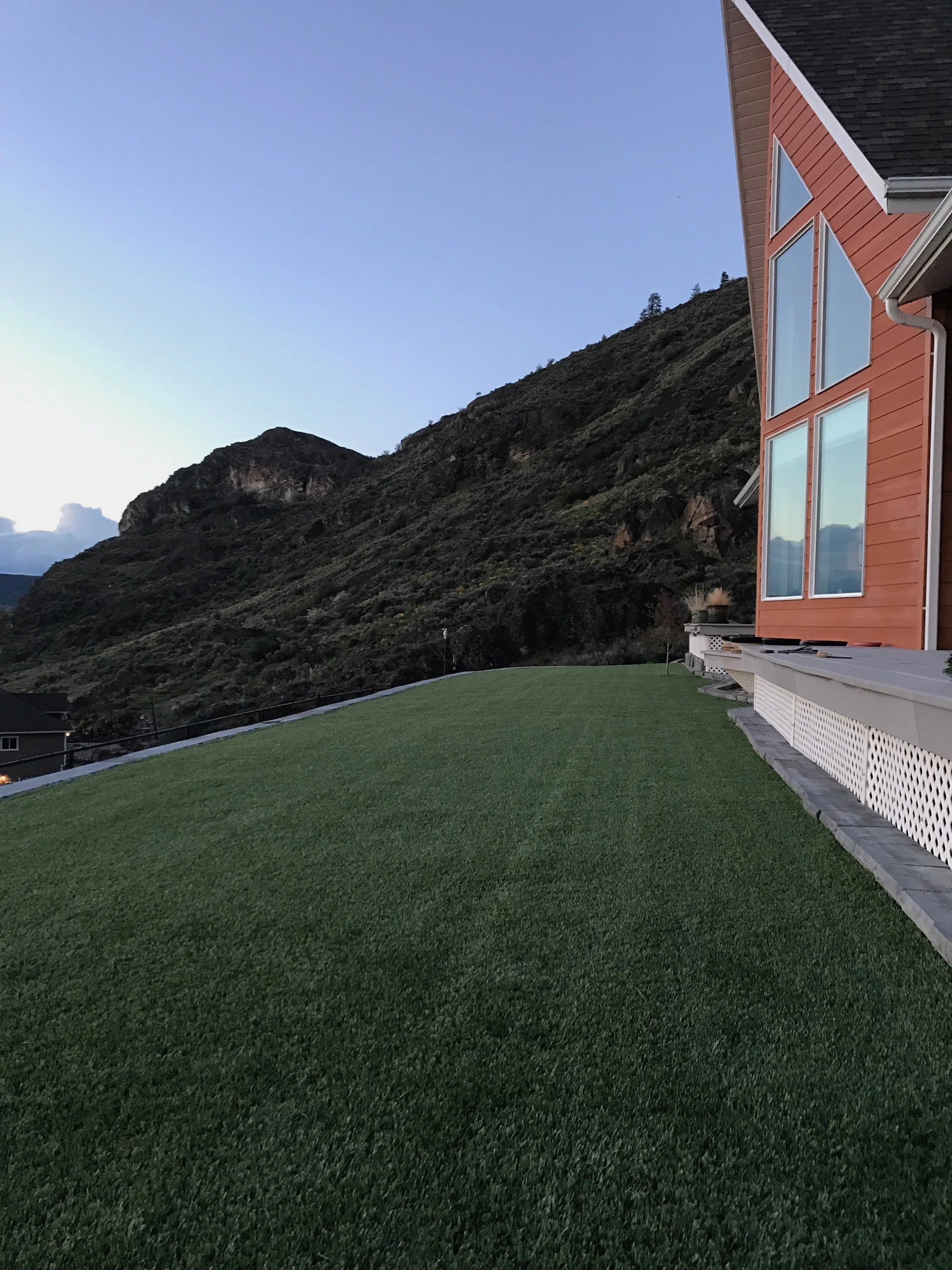 Side view of a house with large windows and orange exterior, with a well-maintained green lawn and mountains in the background during sunset.