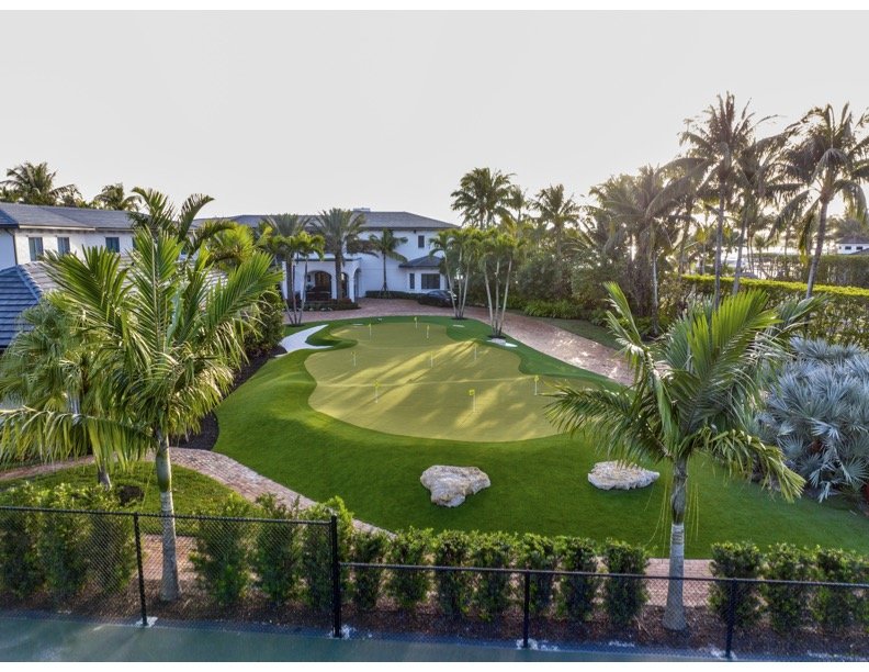 A private mini golf course with a putting green, surrounded by tropical palm trees and lush greenery, located near residential houses.