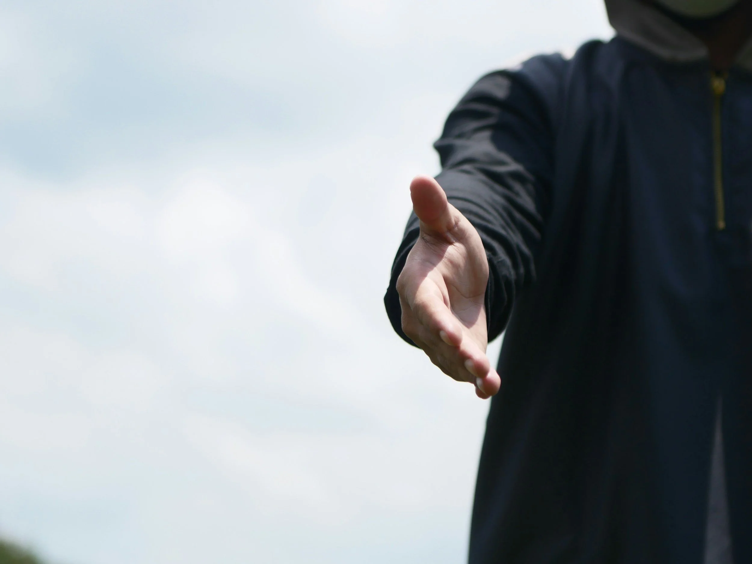 A person in a dark jacket reaching out with their hand against a cloudy sky background.