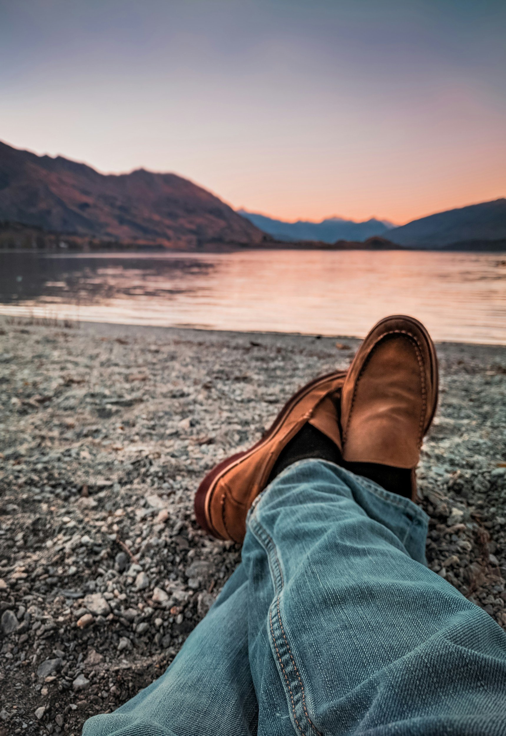 Person relaxing on a rocky beach with boots, jeans, and a scenic lake view surrounded by mountains at dusk.