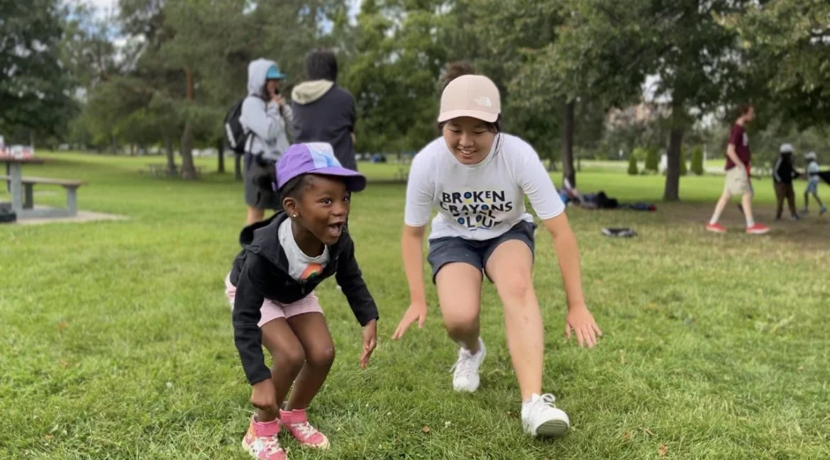 Two young girls are playing on grass in a park, laughing and crouching down. One girl is wearing a black jacket, pink shorts, pink shoes, and a purple cap. The other girl is wearing a white t-shirt with colorful writing, gray shorts, and a beige cap. Other children and adults are visible in the background.