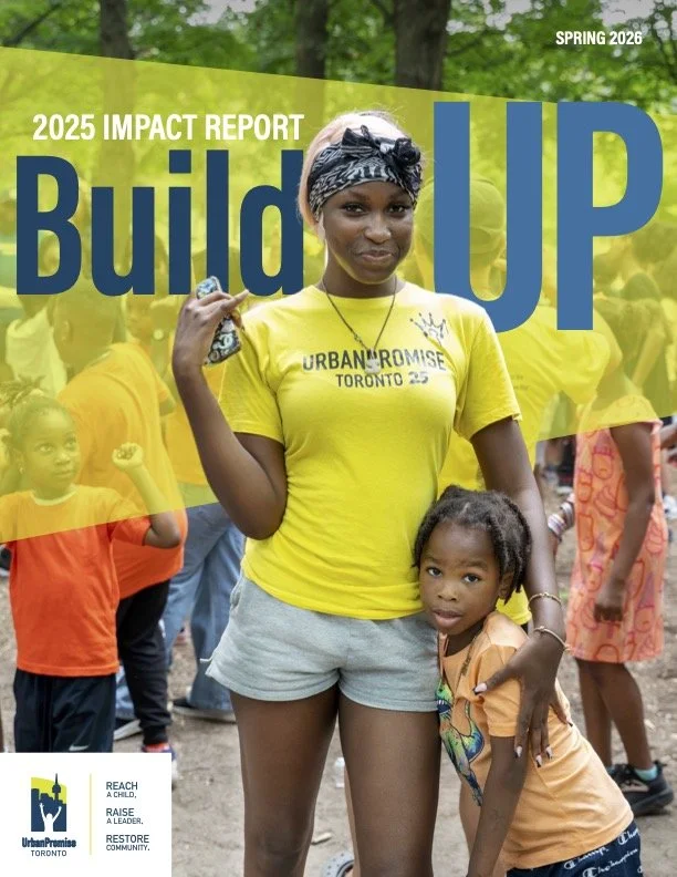 Cover of the 2025 impact report titled Build Up, spring 2026, featuring a woman wearing a yellow shirt and gray shorts, holding a small object, with children and other people in the background, at an outdoor community event.