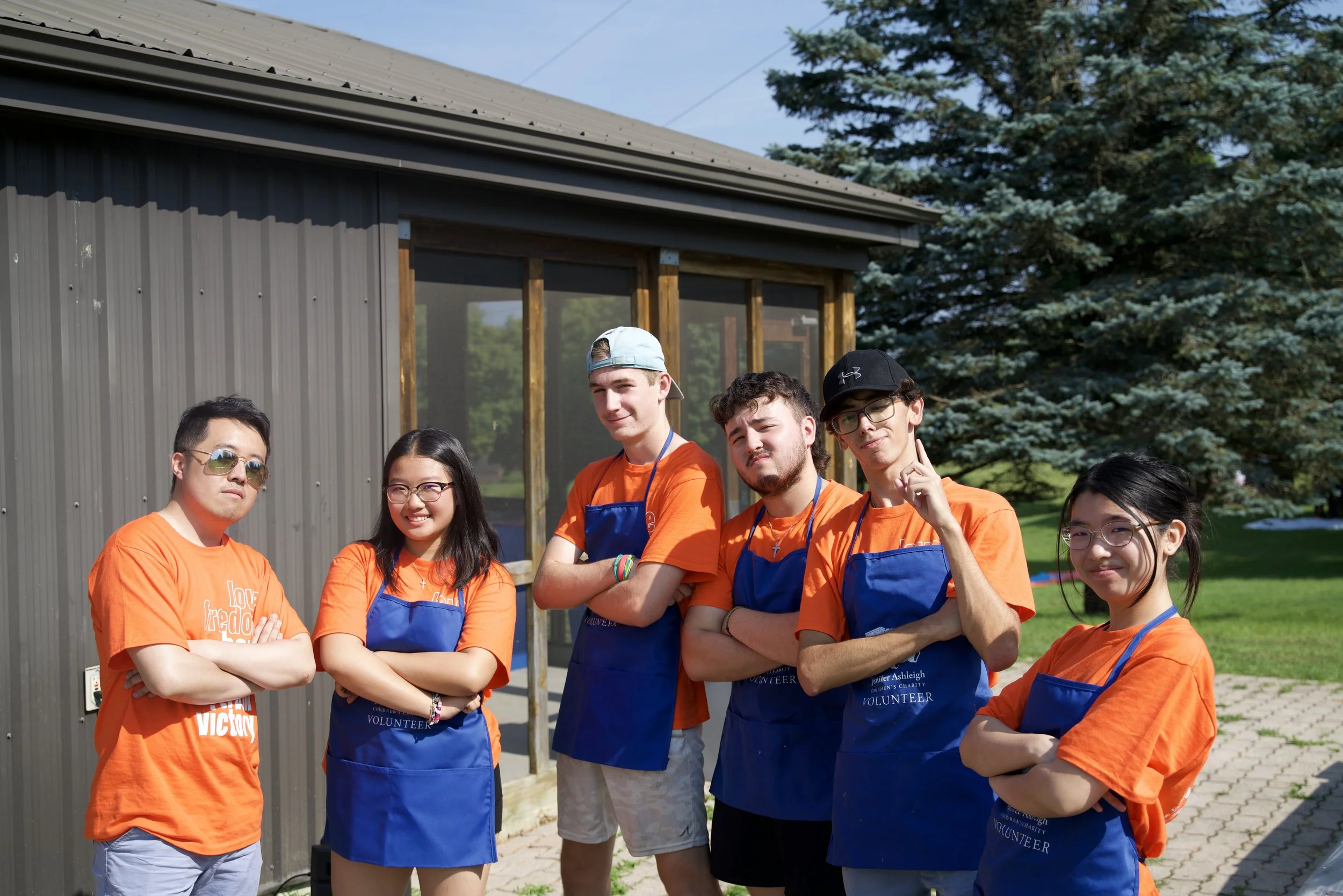 Group of young volunteers wearing bright orange shirts and blue aprons, standing outdoors in front of a grey building with trees in the background.