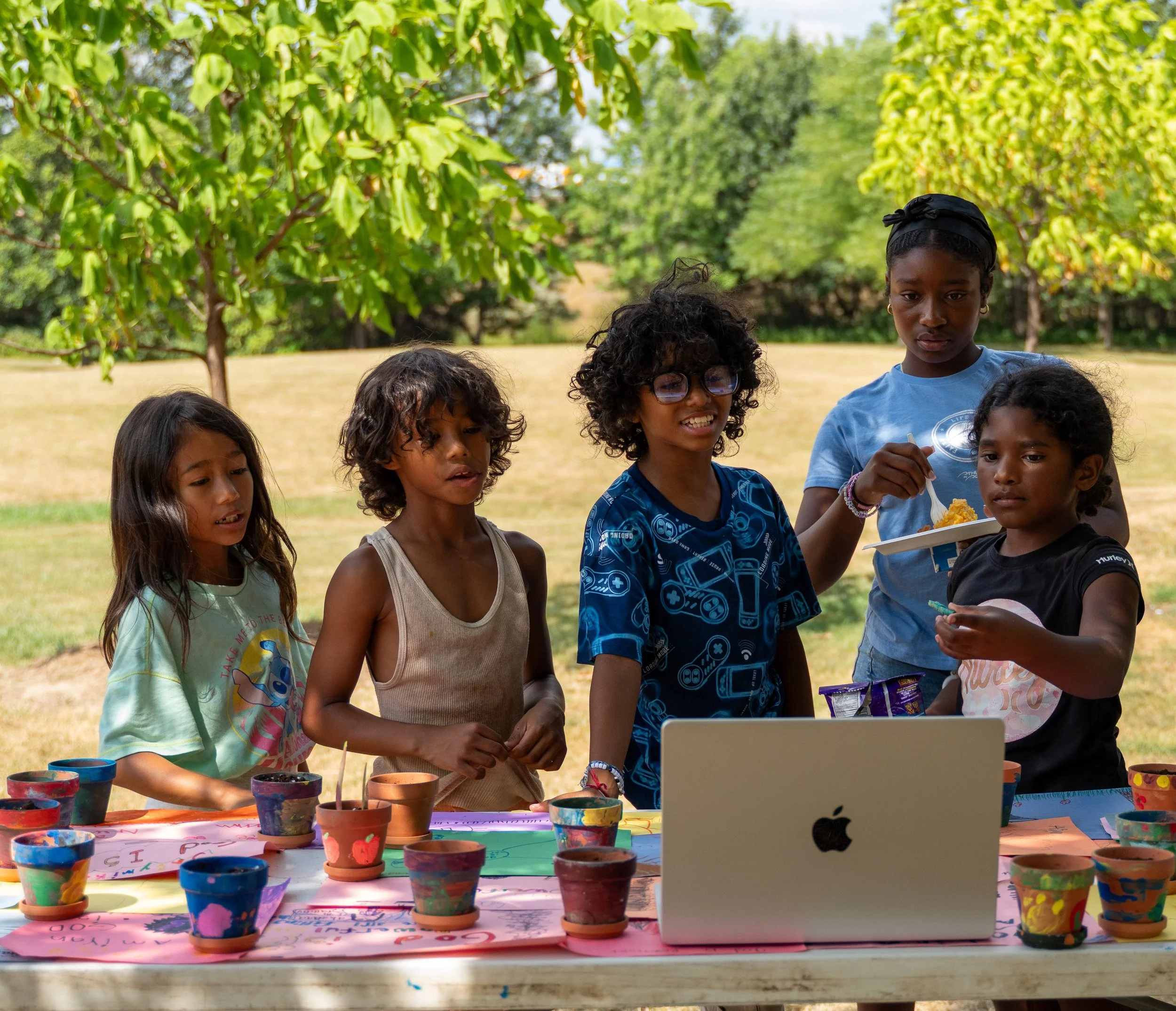 Group of children gathered around a table outdoors, participating in an art activity with colored pots and paint, with a woman in a blue shirt nearby, in a park setting with trees and grass.