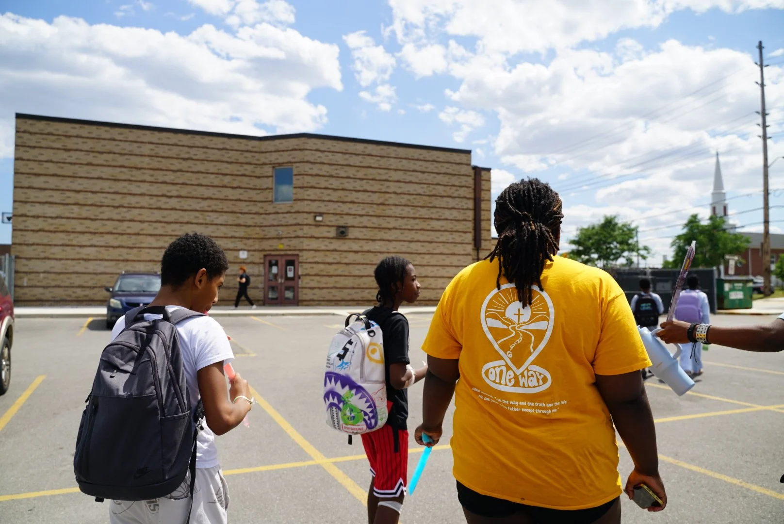 Group of people, including children and an adult, standing in a parking lot outside a building under a partly cloudy sky.