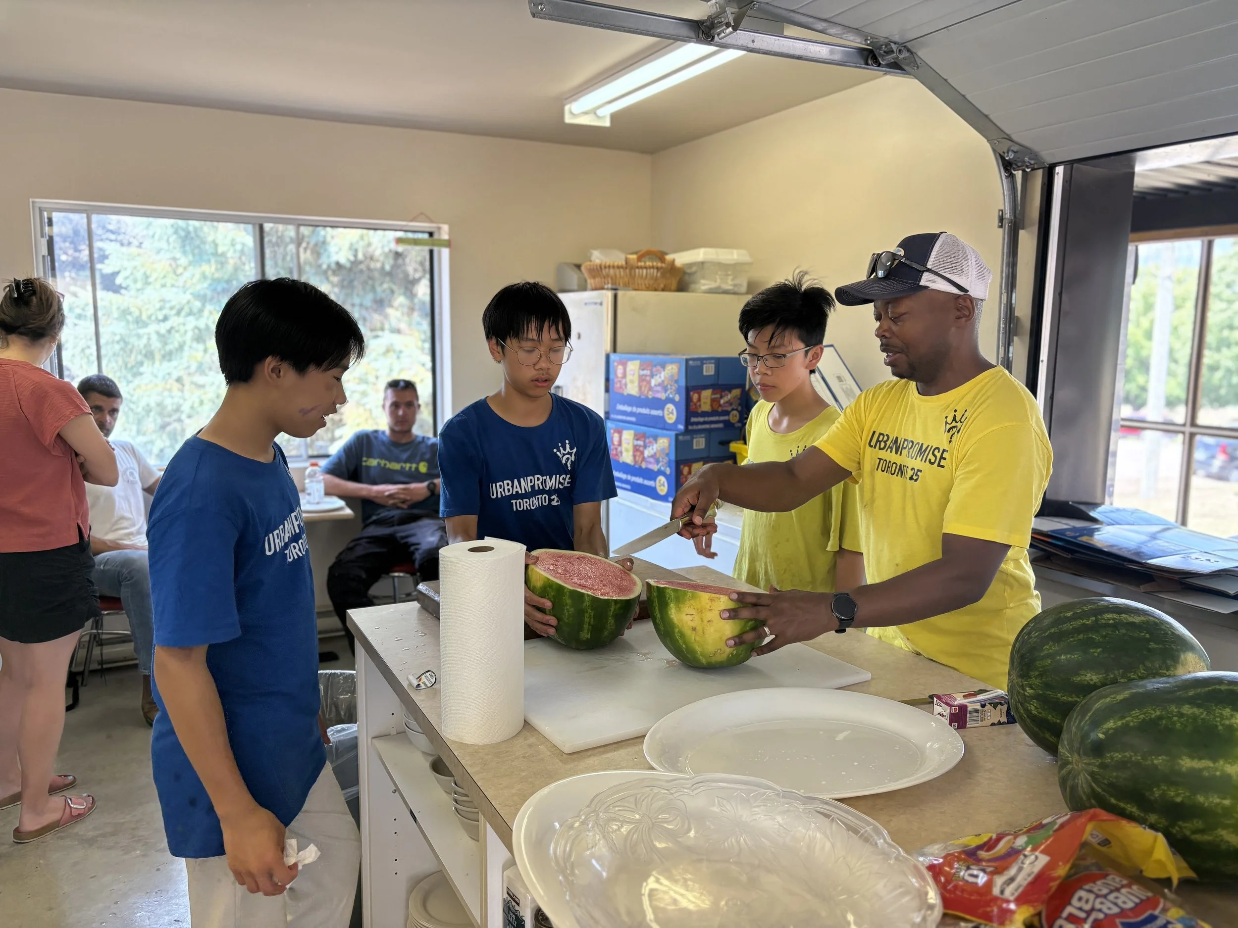 People at a community event or workshop, including a man in a yellow shirt demonstrating or discussing watermelons with a group of children, inside a room with large windows and supplies on the table.