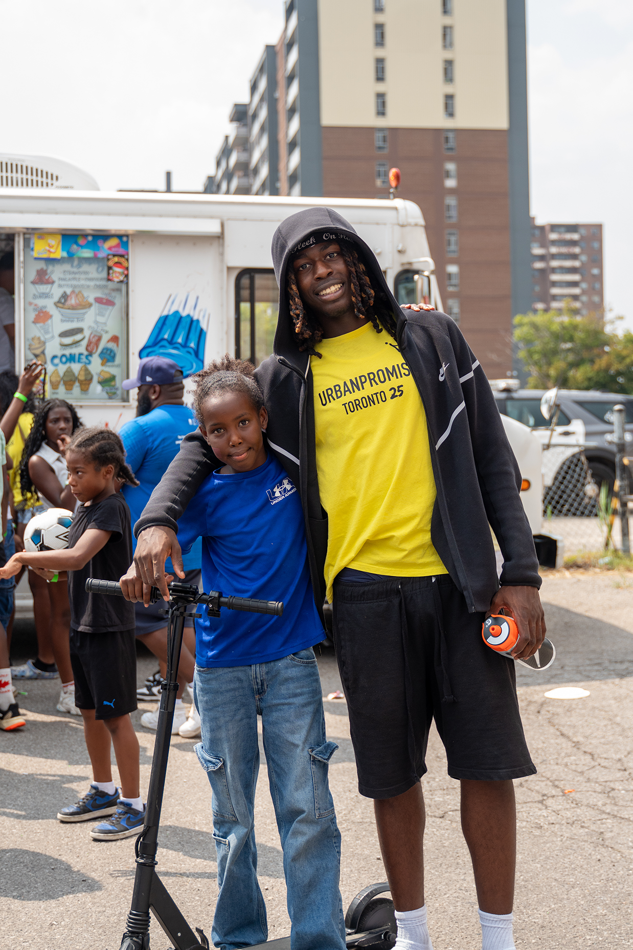 A young man and a girl pose together at an outdoor event, with the young man holding a speaker and a water toy, wearing casual sportswear, and the girl holding a scooter, with a food truck and other children in the background.