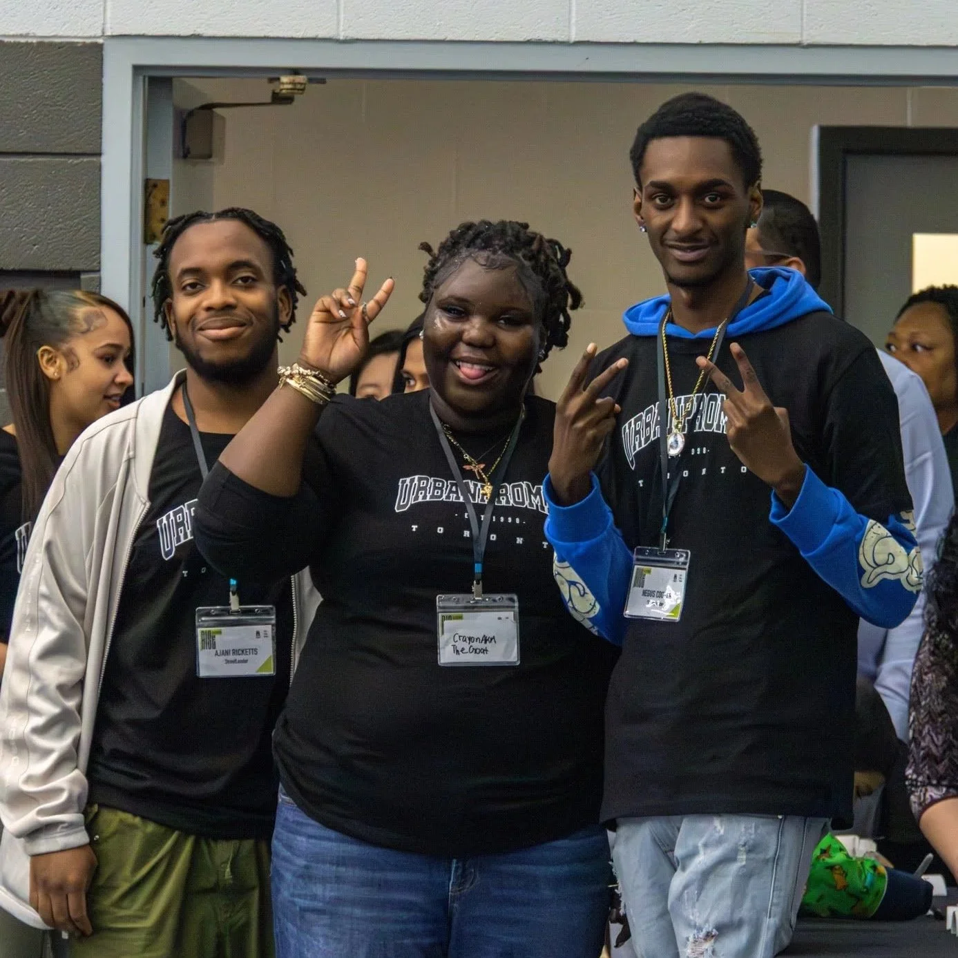 Group of diverse young people at a conference, smiling and making peace signs, wearing black T-shirts with "Urban