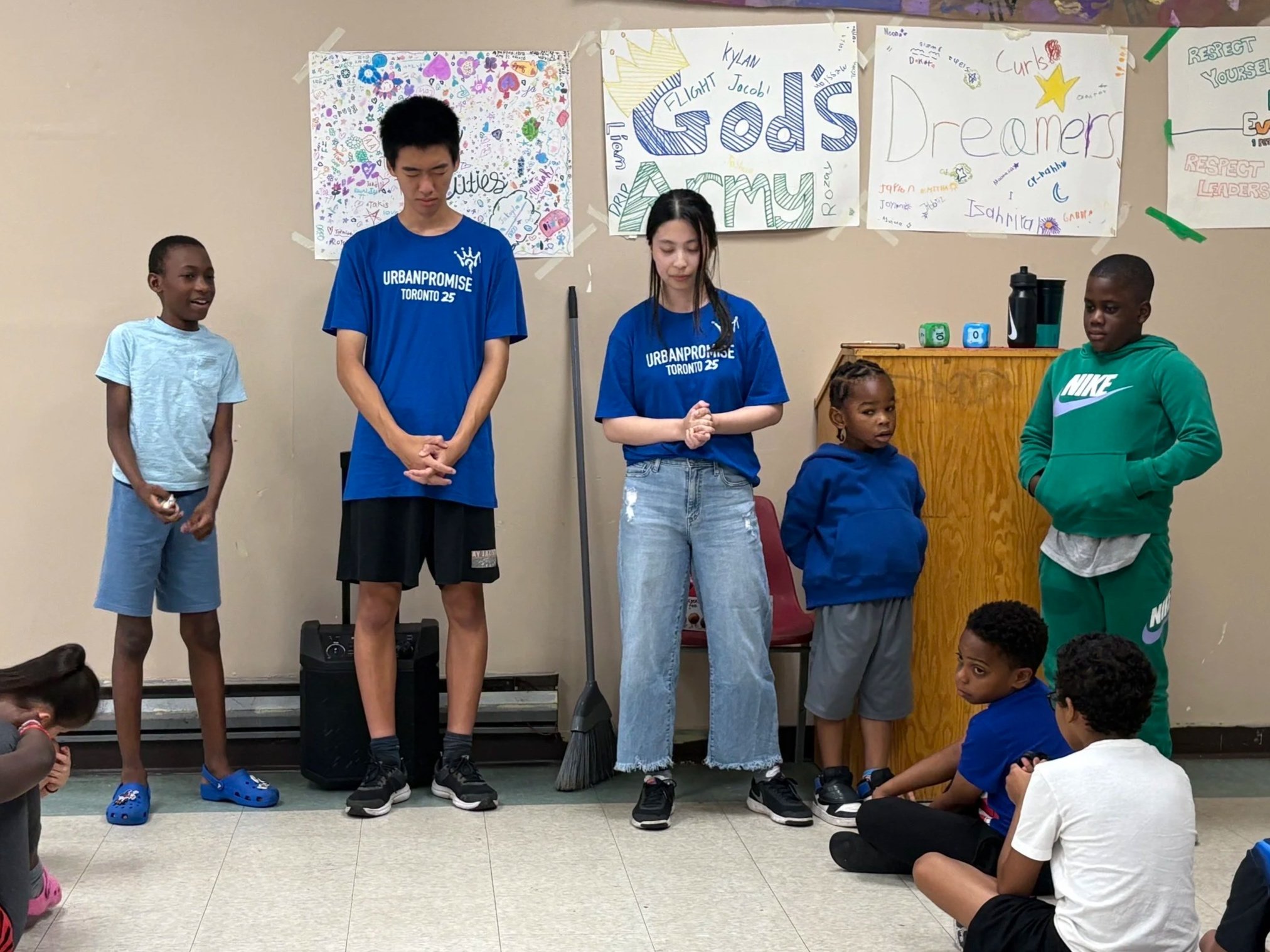A group of children and teenagers standing and sitting indoors near decorated posters on the wall, participating in an activity or presentation.