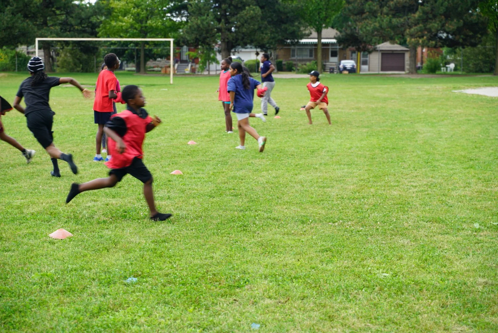 Children playing and running on a grassy field, possibly during a sports practice or game, with small orange cones set up as markers.
